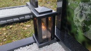 Гранитная прямоугольная лампада с бронзовыми стёклами и зажженной свечой на фоне вертикального стеклянного памятника